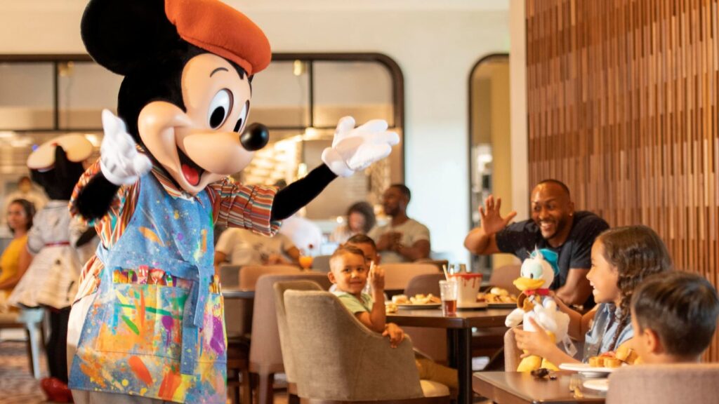  Mickey Mouse vêtu d’une tenue d’artiste colorée, accueillant avec enthousiasme des familles attablées au restaurant Topolino’s Terrace – Flavors of the Riviera, situé au Disney’s Riviera Resort.